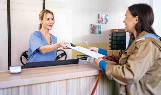 A smiling woman in blue scrubs hands documents to a pet owner at a veterinarian's reception desk.