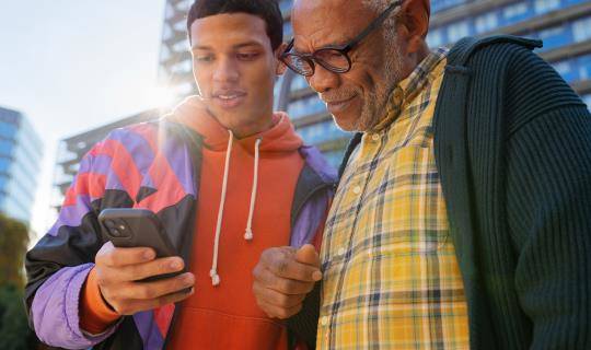 Casual portrait of a young man showing an older man something on his phone, shot on a sunny day in the city