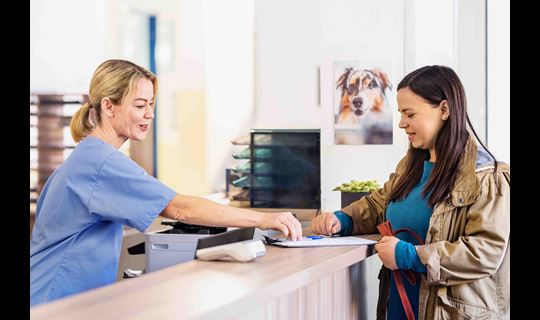 A cheerful woman interacts with a veterinarian receptionist, providing paperwork for her dog's appointment, in a modern clinic setting.