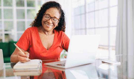 Smiling woman sitting at a table in her home office, writing in a notebook.