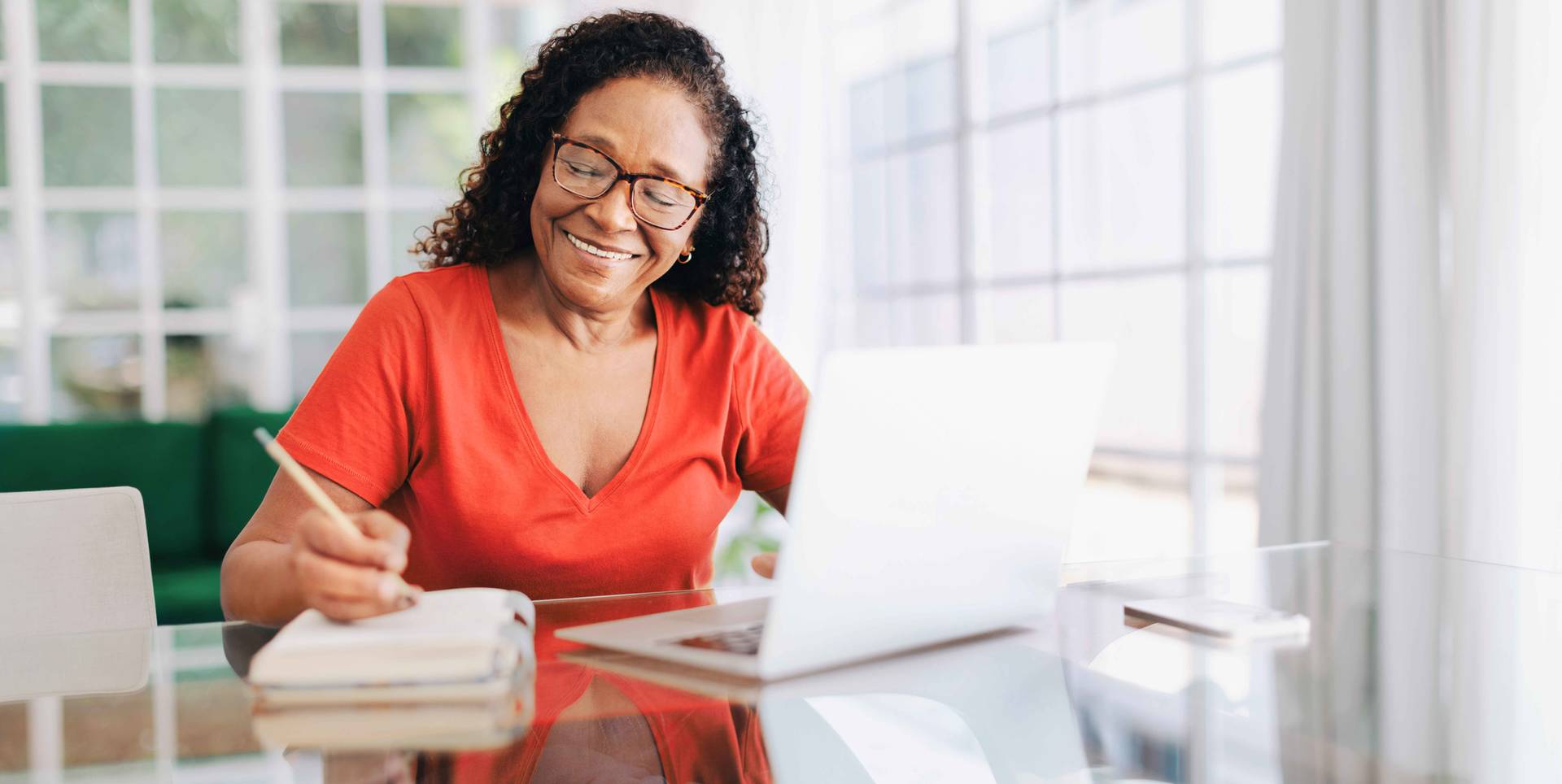 Smiling woman sitting at a table in her home office, writing in a notebook.